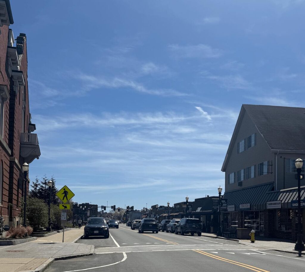Downtown Needham MA looking down Chapel Street with brick storefronts, vintage street lamps, and Needham Center shops on a sunny spring day