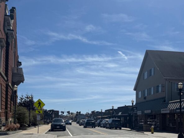 Downtown Needham MA looking down Chapel Street with brick storefronts, vintage street lamps, and Needham Center shops on a sunny spring day